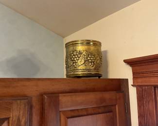 Brass Grapevine Decorative Planter — Heavy brass decorative planter with raised grapevine motif around the body and banded rim and base. Solid metal construction with a warm aged patina and surface wear consistent with display use; a few small dings and light verdigris in crevices but no major dents. Stands on three small feet and works as a planter, umbrella/hat stand cover, or decorative cachepot; sturdy, substantial piece that displays well on a shelf or tabletop. Measurements are approximate and provided for planning removal.