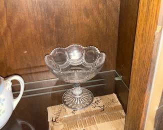 Pressed Glass Compote Dish — Clear pressed glass compote dish with scalloped bowl and pedestal base, ideal for displaying fruit or desserts. The piece features molded decorative panels around the bowl and a starburst pattern on the round foot, finished with a flared scalloped rim. Measures approximately 8 inches tall with a bowl diameter near 6 inches, a lightweight but stable decorative serving piece. In good condition with no visible chips or cracks; minor surface wear consistent with normal use.