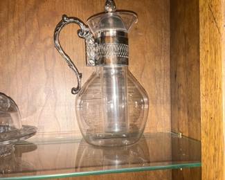 Glass Pitcher with Silver-Plated Collar — Clear glass pitcher featuring a rounded body, flared spout, and ornate silver-plated collar and handle for an elegant serving presentation. The collar has an embossed lattice/leaf motif and the handle is scroll-shaped with decorative detailing; glass body is plain with faint horizontal mold lines. Overall compact size suitable for tabletop use or display in a cabinet; lightweight and easy to handle. In good vintage condition with no visible chips or cracks in the glass, silver-plated mount shows light surface tarnish consistent with age.