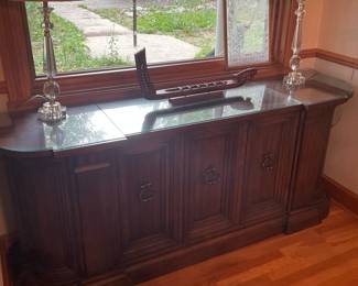 Wooden Four-Door Sideboard Buffet — Solid-looking four-door wooden sideboard buffet with decorative raised panels and metal ring-pull hardware, topped with a fitted glass protector. The piece features a curved front corners and molded base that give it a traditional, formal look suitable for a dining room or entry. Overall footprint is substantial (measurements provided) and includes three or four interior compartments likely useful for storage and serving pieces. Good used condition with light surface wear on top and edges; glass top appears intact and removable.