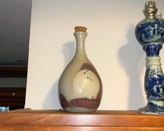 Handcrafted Glazed Stoneware Bottle — A mid-sized handcrafted glazed stoneware bottle with a narrow neck and cork stopper, finished in muted cream, brown and rust tones with abstract landscape-style brushwork. The piece shows handmade character with kiln speckling and glaze pooling at the base; cork top is original-worn but intact. Decorative and sturdy, it works well as a display vessel or accent piece on a shelf or mantel. Good overall condition with light surface wear consistent with age and display; approx. dimensions provided below.