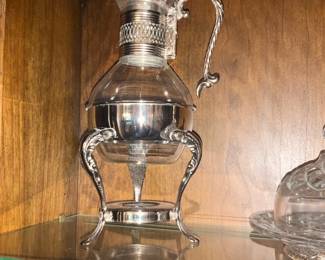Silver Plated Claret Pitcher with Stand — Decorative silver-plated claret pitcher with ornate handle sits on a matching three-legged stand, perfect for display or serving. The set combines pressed glass and silver-plated metal with scrolled, leafy detailing on the handle and legs and a removable glass liner. Overall footprint is moderate and suitable for a china cabinet or sideboard; measurements provided below are estimated. Good estate condition with light surface scratches and expected tarnish on the plating; glass liner appears intact with no visible chips.