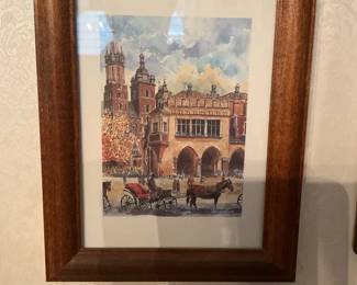 Framed European Watercolor Print — Framed watercolor-style print depicting a European city scene with horse-drawn carriages and historic architecture. Presented in a medium brown wood frame with a white mat; appears ready to hang and suited for entryway or living room display. Overall framed dimensions estimated for wall placement and easy handling. Frame and print show light wear consistent with normal display; glass intact with no visible cracks.