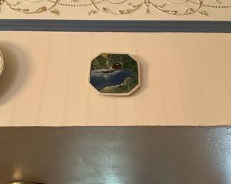 Small Scenic Octagonal Wall Plate — Hand-painted small octagonal wall plate featuring a lakeside scene with a cabin and trees. The ceramic plate has muted greens and blues with a white rim and appears intended as decorative wall art. Plate measures approximately 6 in across and is lightweight for hanging; perfect for a gallery wall or cottage decor. In good condition with no visible chips or cracks from the photo; sold as-is and priced to move at an estate sale.