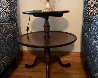 Two-Tier Mahogany Accent Table — Two-tier mahogany-finish accent table with round tiers and turned central column sitting on a tripod base. The smaller top tier overhangs and the lower tier provides additional display surface; overall construction appears medium-quality with a warm brown finish. Shows light surface wear and a few small scuffs consistent with normal use but no major damage or structural issues visible. Compact footprint works well as a side table or lamp table in a living room or bedroom.