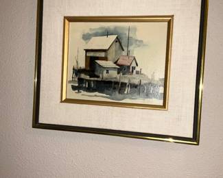 Framed Coastal Watercolor Print — Framed watercolor-style coastal scene depicting dockside buildings with muted blues, greys and a red roof accent. The image is double-matted with a linen-look outer mat and a gold inner frame, all within a slim gold metal outer frame; glazing appears intact. Unframed artwork visible area about 10 x 7 inches (estimate), overall framed piece is lightweight and ready to hang; good decorative condition with minor surface scuffs to frame consistent with normal use.