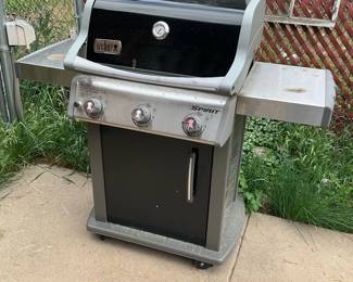 Weber Spirit 3-Burner Gas Grill — Weber Spirit three-burner propane gas grill with side shelves and lower cabinet for a tank. The unit features a black hood with integrated thermometer, stainless-steel control panel with three knobs, and foldable side tables for prep. Overall footprint is moderate and it’s suitable for a patio or deck; measurements listed are approximate. Grill shows exterior wear, surface rust and dirt consistent with outdoor use but appears structurally intact; unknown working condition — propane tank and ignition not included.