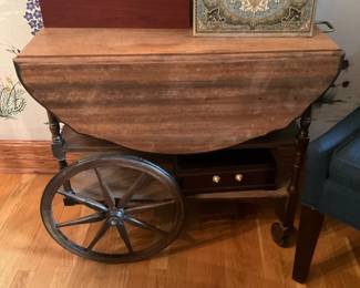 Wooden Drop-Leaf Tea Cart — Rustic wooden drop-leaf tea cart with large wagon-style wheels and turned legs, offering a charming farmhouse look. The cart is constructed of stained/patinaed wood with two large drop leaves, a lower shelf and brass caster on one leg for mobility. Overall footprint is compact and useful as a serving cart or accent table in an entry or living room. Shows wear consistent with age including surface scuffs, patina and some finish loss but is sturdy and functional.