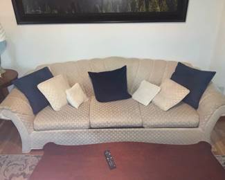 Beige Upholstered Three-Seat Sofa — Three-seat upholstered sofa in a beige patterned woven fabric with serpentine scalloped back and rolled arms. Cushioned bench-style seat is divided into three removable cushions and comes with five assorted throw pillows (two navy, three beige). Construction appears to be hardwood frame with standard foam padding; fabric shows light overall wear and minor soiling consistent with normal use but no obvious tears or structural damage visible in photos. Measures medium-sized for living rooms and would suit traditional or transitional decor.