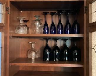 Wood Glassware Cabinet with Shelves — Solid wood cabinet holding an assortment of stemware on two adjustable shelves. The cabinet features warm medium-brown wood with two interior shelves currently styled with clear and cobalt blue wine glasses; hinged doors with leaded glass panels are partially visible. Overall footprint is moderate and suitable for dining room or kitchen storage; shelves appear adjustable to accommodate taller glassware. Cabinet shows light surface wear and scuffs consistent with normal use but is structurally sound and ready for continued use.