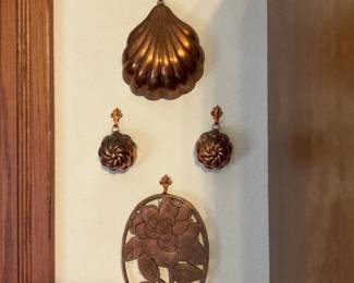 Copper Hanging Molds Set — Decorative set of three copper hanging molds and a carved copper-tone floral plaque shown mounted on a wall. The largest mold is a scallop-shell shape with two smaller spiral molds below, all with small fleur-de-lis style hangers; the oval cutout floral plaque appears to be stamped or cast metal with a rose motif. Warm aged copper patina with light surface wear and tarnish consistent with age; no obvious breaks or dents visible in photos. Useful as kitchen or wall decor, total display height about 20 inches for the group with individual pieces removable for cleaning or reuse.