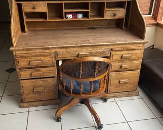 Oak Roll-Top Desk — Solid oak roll-top desk with multiple small cubbies, drawers and a drop-front workspace that rises to reveal pigeonholes and two small drawers. The finish shows even patina and honest surface wear consistent with regular use, with some scuffs on drawer fronts and edge wear at the base. Sturdy construction with traditional pulls and a removable/slide roll panel; a swivel wooden chair with caned back is pictured but sold separately unless requested. Measures roughly 48 inches wide by 30 inches tall at the roll top and 24 inches deep.