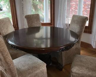 Carved Pedestal Round Dining Table — Solid wood round dining table with a carved central pedestal and scrolled tripod base, shown with six slipcovered high-back chairs. The rich dark finish and carved apron/leg details give it a traditional, formal look; pedestal construction allows generous legroom. Tabletop shows light surface wear and some small finish scuffs consistent with normal use; base carving and chair slipcovers are intact though chairs show light fabric wear. Measures roughly 48 inches diameter and standard dining height; sturdy, heavy construction will require two people to move.