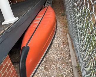 Orange Recreational Canoe — Bright orange recreational canoe suitable for lakes and calm rivers, shown stored upside-down beside a porch. Fiberglass or polyethylene hull with black gunwales and aluminum/metal end caps visible; light scuffs and surface wear from outdoor storage but no major cracks seen. Overall length appears to be a standard canoe size appropriate for 2–3 paddlers — compact enough for roof or trailer transport. Being stored outdoors has left cosmetic wear and dirt; recommend cleaning and close inspection of hull for any stress fractures before use.