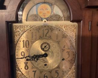 Howard Miller Grandfather Clock — Tall floor-standing grandfather clock featuring a carved bonnet top and brass dial with moon phase complication. The solid wood case houses visible brass weights and lyre-style pendulum behind a full-length glass door, finished in medium brown. Measures approximately 84 inches high, 20 inches wide, and 14 inches deep and is substantial but movable by two people. In good working cosmetic condition with light surface wear to finish consistent with age; clock appears intact though movement function should be verified by buyer.