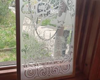 Etched Glass Scenic Panel in Frame — Decorative framed etched glass panel featuring an oval pastoral scene with trees, cottages and birds surrounded by scrollwork borders. The piece is set in a simple wooden frame and appears designed for display on a window ledge or hung on a wall to catch light. Estimated overall size fits a small-to-medium window—see dimensions for specifics; lightweight and easy to move. In good vintage condition with no visible cracks in the glass and only minor scuffs to the wood frame from normal use.