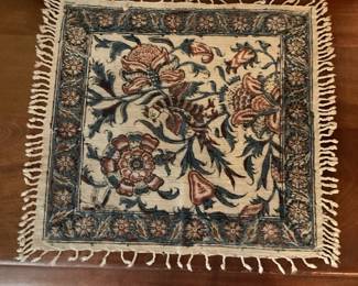 Floral Woven Table Runner Mat — Handwoven-looking square mat with a traditional floral/foliate border and central bouquet motif in muted blue, rust and cream tones. The piece has short fringe on two sides and shows light surface wear and slight age-related discoloration consistent with use; no major stains or holes visible. Use as a vanity or table accent, under a vase or lamp — lightweight and easy to place. Measures small and decorative; see dimensions for exact size.