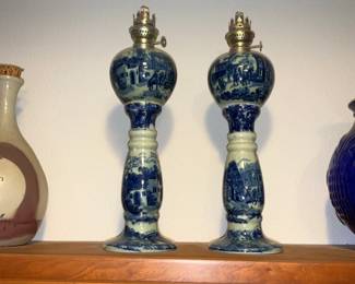 Blue & White Porcelain Oil Lamps — A matched pair of tall blue-and-white porcelain oil lamps with painted village and landscape scenes on baluster bodies and bulbous tops. Each has original brass burner fittings; decorative cobalt designs run the full height with a flared circular base. They show light surface wear and small glaze crazing consistent with age but no obvious chips or large cracks visible in photos. Overall height is substantial and they make a striking decorative pair for mantel or shelf display.