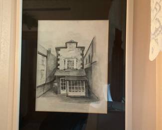 Framed Charcoal Architectural Sketch — Framed black-and-white architectural charcoal sketch depicting a narrow street façade with a shopfront and upper windows. The drawing is matted with a wide black mat and housed in a simple painted wood frame with glass. Overall framed size is estimated for wall display and easy hanging; frame is lightweight but adds protection for the paper artwork. In good condition with light surface reflections on the glass seen in photos and no visible tears or major staining to the artwork.