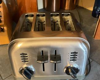 4-Slot Stainless Toaster — Four-slot stainless steel toaster with dual controls and function buttons labeled bagel, defrost, reheat and cancel. Heavy-duty metal housing and separate levers/dials for each two-slot pair allow independent browning settings. Shows moderate surface wear and light interior crumbs from use but appears intact with all controls present; cord visible. Overall a practical kitchen appliance for households needing extra-slot capacity.