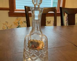 Cut Crystal Decanter with Label — Elegant cut crystal decanter featuring a faceted stopper and deep diamond-cut pattern on the bulbous body. It includes a decorative gilt bottle label on a chain engraved "BRANDY" (visible) adding period charm. Measures approximately 11.5 inches tall with a 4.5-inch diameter base, making it a standard tabletop decanter for spirits. In good condition with no visible chips or cracks; stopper fits well and gold-tone label shows mild surface wear consistent with age.