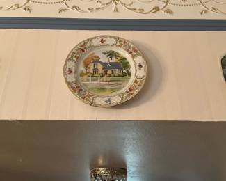 Decorative Scenic Wall Plate — Porcelain decorative wall plate featuring a painted cottage scene with a floral and gilt border. The round plate has detailed hand-painted or transfer-printed imagery and gilt accents around the rim, intended for wall display as an accent. Estimated diameter approx. 10 inches; lightweight and easy to hang. Condition shows a visible hairline crack at the rim (photo) and some wear to gilt and surface typical of age.