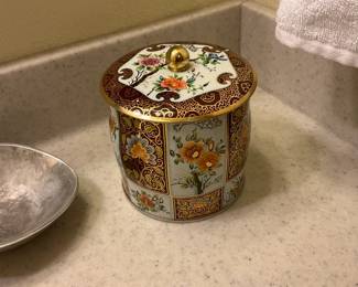 Decorative Floral Covered Tin — Round decorative covered tin with a hand-painted look floral panel design and gilt accents; the lid has a gold-tone knob and detailed scroll borders. The enamel-like painted panels show colorful chrysanthemums and blossoms in warm orange, blue and green tones framed by a deep brown and gold patterned border. Sturdy sheet-metal construction with bright finish; no visible dents or rust from the photos and only light surface wear consistent with normal use. Compact storage jar suitable for bathroom, kitchen counter, or display; see dimensions for overall size.