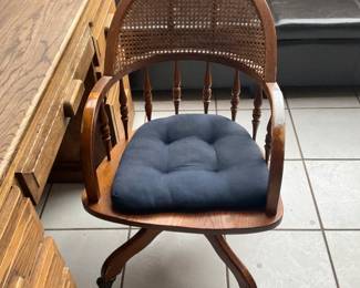 Wooden Caned Swivel Desk Chair — Solid wood swivel desk chair with a rounded caned back, turned spindle supports and a curved X-style base with casters. The original cane back and medium-tone wood finish give it classic traditional character; a tufted navy seat cushion is shown but may be removable. Construction appears sturdy with typical light surface wear and a small area of finish loss on one arm as pictured. Overall a functional, space-saving desk chair that measures compactly for home office or writing desk use.