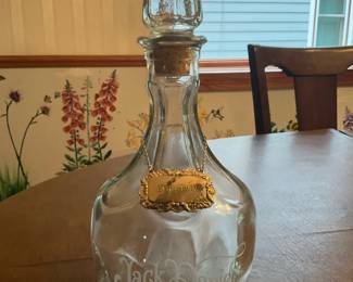 Jack Daniel's Glass Decanter with Tag — Clear glass Jack Daniel's decanter featuring a faceted glass stopper and an ornate gilt metal label hung on a chain reading 'Bourbon'. The bulbous bottle has etched 'Jack Daniel's' script on the front and is made of thick clear glass with a traditional spirits decanter form. Measures approximately 12 in tall to top of stopper, 6.5 in wide at the shoulder, and 4.5 in deep; a sturdy, empty decanter ideal for display or tabletop use. In good estate condition with light surface wear and a few small scuffs consistent with age, no visible chips or cracks in photos.