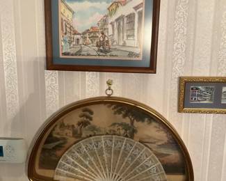 Decorative Framed Lace Hand Fan — Framed decorative lace hand fan displayed in a semicircular gold frame with a scenic painted background. The folding fan features delicate white lace and carved sticks, showcased behind glass for wall display. Overall wall-hung piece measures approximately 18 inches wide and 10 inches tall and is lightweight enough for standard wall mounting. In good vintage condition with minor age-typical discoloration to the backing and light wear to the lace; glass and frame appear intact.
