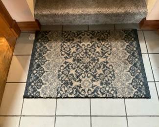 Gray Patterned Indoor Accent Rug — Neutral gray patterned indoor accent rug featuring a faded medallion and floral border motif that works well in entryways or small living spaces. Low-pile synthetic construction with a simple bound edge and non-slip backing visible at the corner; colors are gray and charcoal with a light distressed look. Estimated footprint fits a small doorway or foyer — see dimensions below for exact measurements; lightweight and easy to move. Rug is in good condition with normal light wear from use and no visible stains or rips.