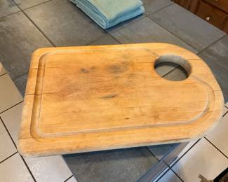 Thick Wood Carving Cutting Board — Large, thick hardwood cutting board with a carved juice groove and integrated hand hole for easy carrying. Solid construction shows knife marks, surface staining, and some edge wear consistent with regular kitchen use but remains structurally sound. Ideal as a heavy-duty prep board or butcher-block top; will benefit from sanding and oiling to refresh the surface. Overall a practical, well-built board that measures roughly 20 x 14 inches and is easy to handle thanks to the carved grip.