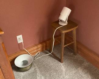 Small Wooden Stool — Simple solid-wood stool with a square saddle-style seat and four straight legs with stretchers; shown with a small white spotlight resting on top. Construction appears to be light to medium hardwood with a natural finish and visible wear at the seat edges and legs consistent with normal use. Compact and lightweight for easy moving — useful as a plant stand, side seat, or display stand. Overall fair to good condition with scuffs, finish wear, and surface marks as seen in photos.