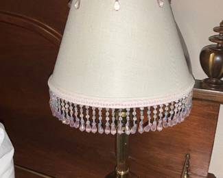 Brass Table Lamp w/ Pink Beaded Shade — Brass table lamp with a tapered fabric shade trimmed in pink beads and dangling pink teardrop prisms. The lamp has a turned brass column and round brass base with a traditional pull-chain socket; the shade features two-tier bead fringe and small bead drops around the top edge. Good overall cosmetic condition with light surface wear and minor tarnish to the brass consistent with normal use; shade appears intact with all fringe present. Compact bedside size and ready to use with a standard bulb.