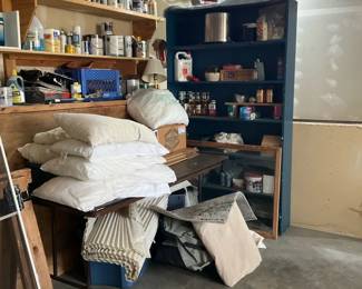 Garage Storage Shelves & Misc Items — A mixed lot of garage storage and household items shown together: a tall painted open shelving unit filled with paint cans, jars and small hardware, a folding table stacked with pillows and fabric/trim, and assorted shelving and containers. Ideal for someone looking to clear out workshop supplies or pick up usable shelving, containers, and home project materials. Overall dimensions estimated for the shelving unit and table are provided; items are used and show typical dust and wear from garage storage. Sold as-is — contents are included but not inventoried and some smaller items may be missing or need testing.