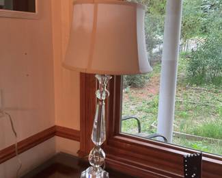 Crystal Glass Table Lamp with Shade — Elegant crystal glass table lamp topped with a neutral beige fabric drum shade, offering classic sparkle and reflective accents for a living room or bedside. The lamp features a multi-faceted crystal column with stacked orb and geometric elements on a solid crystal base, with standard harp and finial hardware. Overall height is approximately 28 inches including the shade; the lamp is easy to place on side tables, dressers, or entry tables. In good working condition with light surface wear on the shade and minor scuffs to the base consistent with normal use.