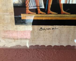 gamal Egyptian Papyrus Print — Hand-signed "gamal" papyrus print depicting a colorful Egyptian funerary scene with hieroglyphs and figures. The print is mounted under clear plastic with scalloped papyrus edges visible; colors remain bright though the papyrus shows age toning and minor edge loss. Signed lower left, it is a decorative reproduction on papyrus typical of tourist or decorative Egyptian-style art. Framed/mounted as shown; condition good with wear to papyrus edges and light surface reflections from the protective covering.