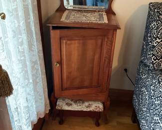 Mahogany Washstand with Mirror — Solid mahogany washstand featuring a shaped mirror backboard, single panel door with brass knob and a matching floral-upholstered footstool. The piece shows warm patinated wood and traditional cabriole legs; mirror has light spotting and the top shows minor surface wear consistent with age. Compact and sturdy, it works well as a bedside cabinet or small hall accent; stool fabric has light discoloration but no visible tears.