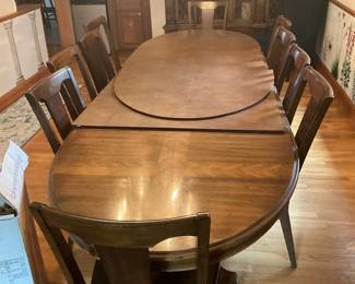 Heritage Grand Manor Oval Dining Set — Solid wood Heritage Grand Manor oval dining table with eight matching chairs, offering a traditional formal dining look. The set is constructed from stained hardwood with a warm medium-brown finish and features an extension leaf (shown separated) to expand seating. Table measures approximately 30 in high, 84 in long (with leaf installed) and 42 in wide; chairs are standard dining height; overall sturdy, suitable for everyday use. Good condition with surface wear, light scratches and finish fading in places consistent with regular use; hardware and joinery appear intact and usable.