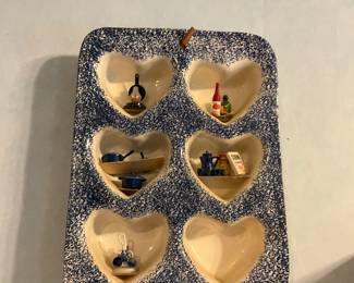 Heart-Shaped Ceramic Wall Shelf — Decorative ceramic wall shelf with six heart-shaped recessed cubbies finished in a blue speckle over cream glaze. Each small niche currently holds miniature kitchenware and collectibles; shelf has a single sawtooth hanger visible at the top for wall mounting. Sturdy glazed pottery construction with rounded corners and a folk-art look — good for displaying small figures, thimbles, or miniatures. Shows light surface wear and a few small glaze imperfections consistent with age but no visible large chips or cracks.