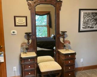 Victorian Carved Walnut Vanity Mirror — Tall Victorian-style carved walnut vanity with an integrated mirror and paired marble-top bedside cabinets. The piece features ornate cresting with finials, carved foliate details and turned candlestick-style supports; both lower cabinets have multiple small drawers with porcelain knobs and marble tops. Construction and joinery appear consistent with late 19th–early 20th century styling; finish shows light surface wear, small scratches and usual age patina but no major structural damage visible. Overall a decorative, substantial bedroom dressing piece that includes the upholstered matching vanity stool shown.