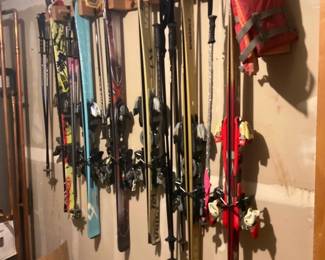 Assorted Downhill Skis with Poles — Lot of assorted downhill alpine skis hanging on a wall rack, offered together with multiple pairs of adjustable ski poles. The group includes several brands and lengths (adults and youth) with integrated bindings visible on most skis; colors range from red and gold to teal and patterned graphics. Overall useful for resale or local pickup; sizes vary but most skis appear in the 150–180 cm range and poles are standard adjustable lengths. Condition is used — some visible scuffs and edge wear typical of regular mountain use, bindings appear intact but are untested and should be inspected and adjusted by a technician before use.