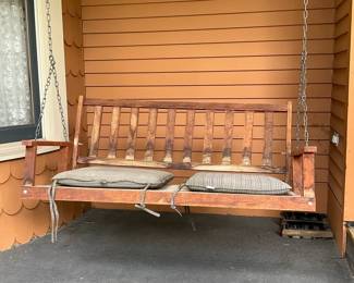 Wooden Porch Swing with Cushions — Solid wooden three-seat porch swing suspended by metal chains, offered with two outdoor seat cushions. Constructed from stained hardwood with slatted back and armrests; shows weathering and sun-fade on the finish. Overall dimensions approximate a typical three-person swing and are suitable for porches or covered patios; chains allow hanging from overhead beam. Condition is fair with surface wear, some discoloration and patina consistent with outdoor use; cushions show light soiling but appear usable.