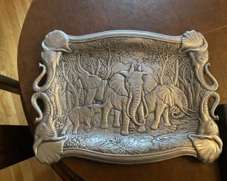 Elephant Relief Decorative Metal Tray — Decorative metal serving/display tray with a detailed elephant family relief scene and molded elephant-head handles at each corner. The tray appears cast from a pewter-toned metal with textured foliage and river details across the interior surface, offering a strong decorative presence for a table or wall. Estimated overall size fits on a small dining table (see dimensions) and it's solid but not overly heavy, making it easy to move or use as a centerpiece. Condition is good with minor surface wear and light handling marks consistent with use; no visible cracks or repairs.