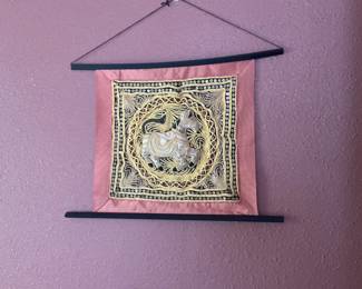 Embroidered Elephant Wall Hanging — Decorative embroidered wall hanging featuring a central elephant motif in gold thread with sequined border on a pink fabric ground, mounted between two black wooden dowels with hanging cord. The textile shows intricate hand-stitched metallic thread work and mirrored sequins typical of South Asian decorative panels and is lightweight for easy display. Good overall condition with no obvious stains or tears visible in photos; minor wear to fabric edges consistent with prior hanging. Overall shallow profile makes it suitable for hallway or living room accent wall.