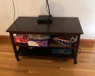 Dark Wood TV Stand Console — Compact dark-stained wood TV stand with two open shelves and simple square legs on small caster feet. The piece is suitable for a media player and board game storage as shown; top surface has minor surface reflections consistent with light use and the shelves show normal shelf wear. Straightforward, neutral design that fits small living rooms or apartments; cords may be routed behind the unit. Overall good condition with only light cosmetic wear to finish.