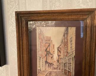 Framed Street Scene Watercolor Print — Charming framed watercolor print depicting a narrow historic street with timbered buildings and cobblestones. The print is double-matted in burgundy and housed in a medium brown wood frame with glass. Overall framed piece is a manageable wall accent suitable for a hallway or living room; measurements provided below. Condition is good with intact glass and frame showing minor surface wear consistent with normal use. No visible artist signature or maker's label legible in the photos.