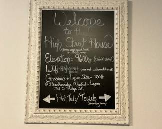 Ornate White Framed Chalkboard — Decorative wall chalkboard with an ornate painted wood frame and black writable surface. Frame shows carved floral and bead details with distressed white paint revealing small areas of underlying color; the chalkboard surface is intact and currently written on as shown. Measures a manageable size for hallway or kitchen message use and is lightweight enough to hang on a single wall anchor. Condition is good with cosmetic paint distressing consistent with decorative use.