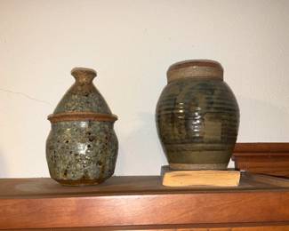 Stoneware Lidded Pottery Jars — A pair of handcrafted stoneware jars with rustic salt-glazed finishes and earthy green-brown tones. One jar is lidded with a flared top and decorative drilled pattern; the other is a stout storage jar with a slightly tapered shoulder and wide mouth. Both show maker-style wheel throwing and natural ash glaze variation; each has light wear consistent with display but no obvious cracks visible in the photos. Useful as decorative vessels or for dry storage; overall compact display pieces.