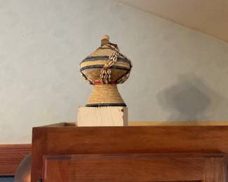 Woven African Basket with Shells — Handwoven decorative basket with a bulbous lidded form and braided cowrie-shell trim. Natural grass/raffia construction shows dyed horizontal bands in blue and yellow with a red accent near the neck; the top finial and lid are intact. Suitable as a shelf accent or for light storage; shows gentle wear consistent with age and use but no obvious breaks in the weaving. Measures small and lightweight—easy to display on a shelf or mantel.